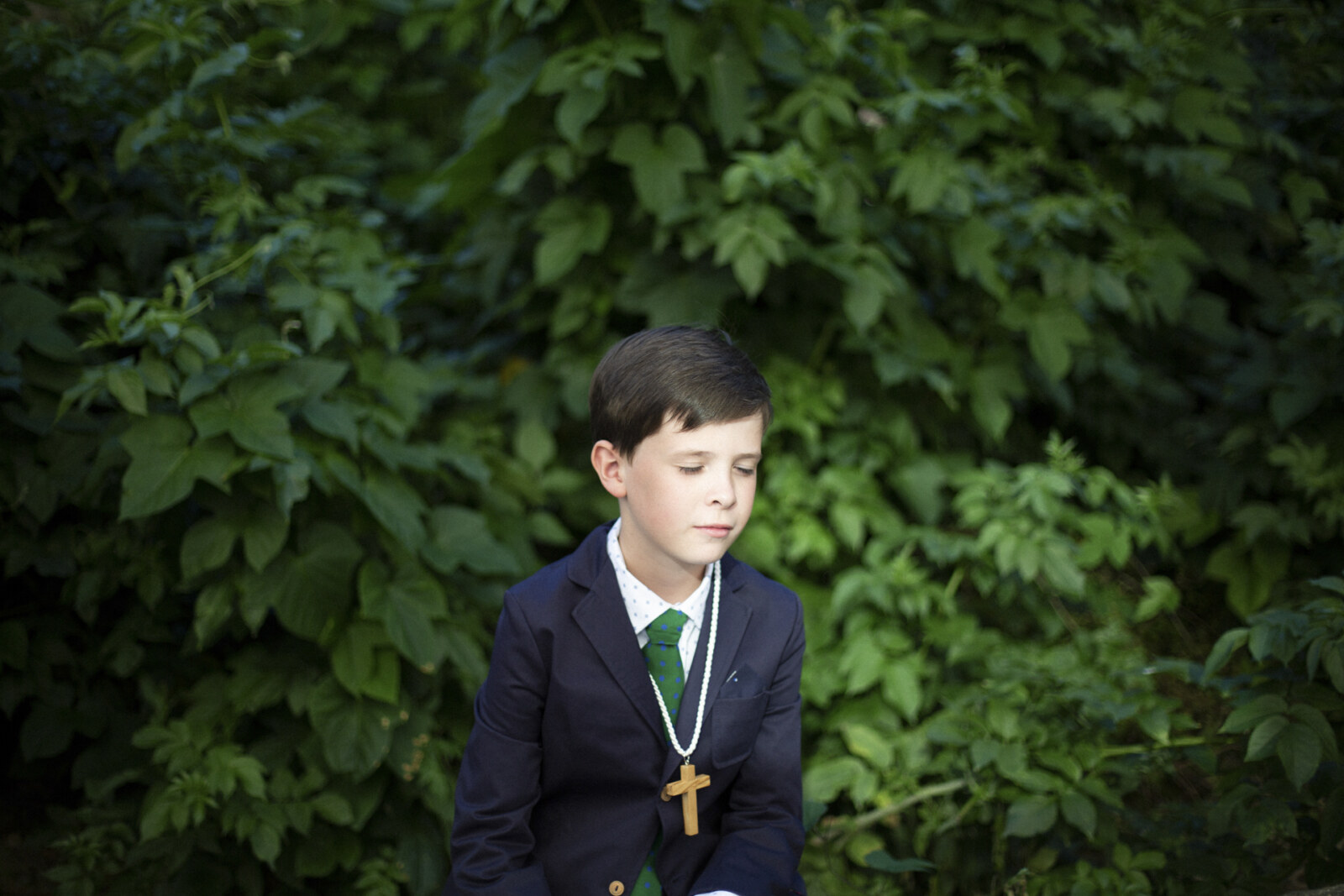 Niño de comunión con corbata verde frente a un seto