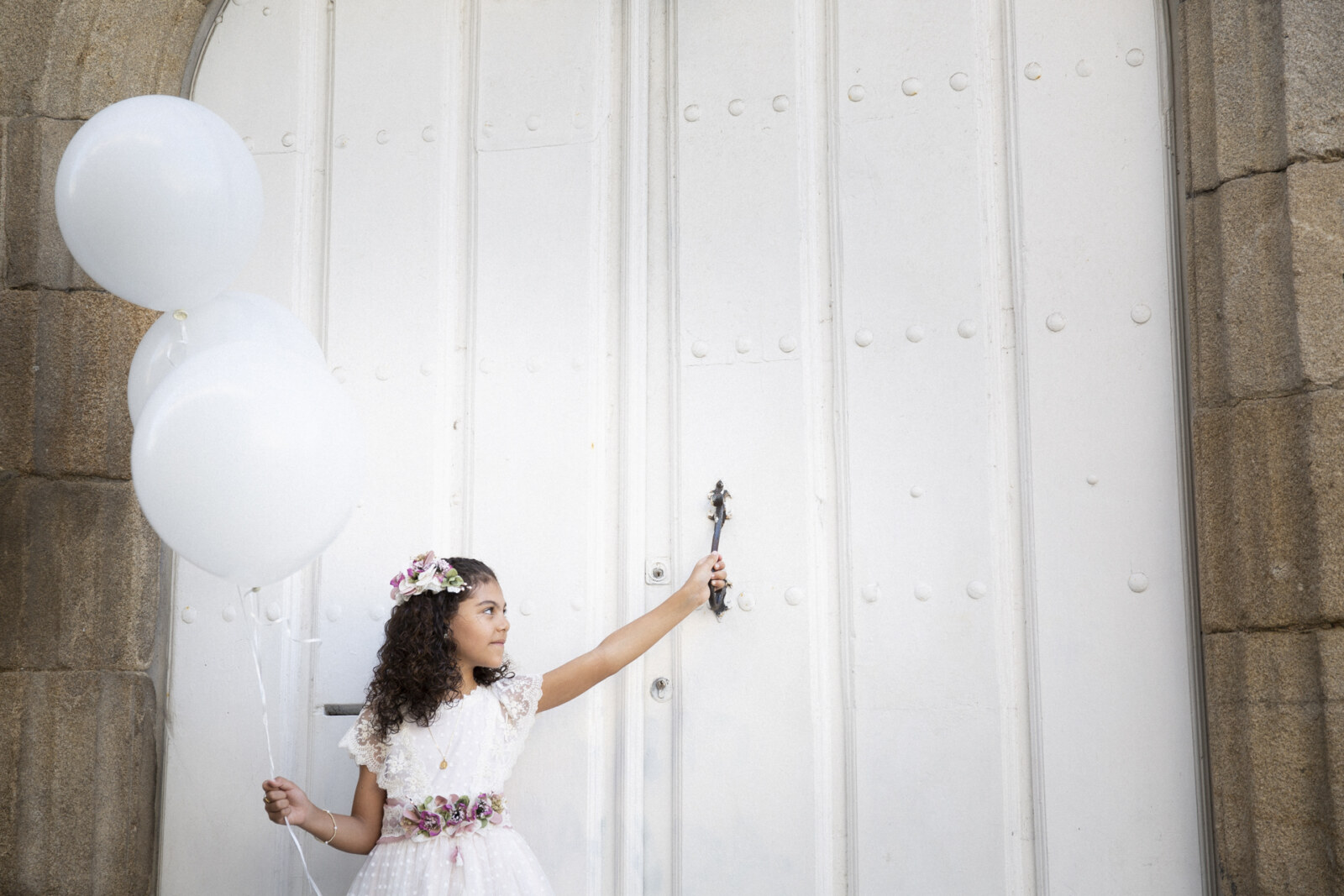 Niña de comunión con globos blancos junto a una pared