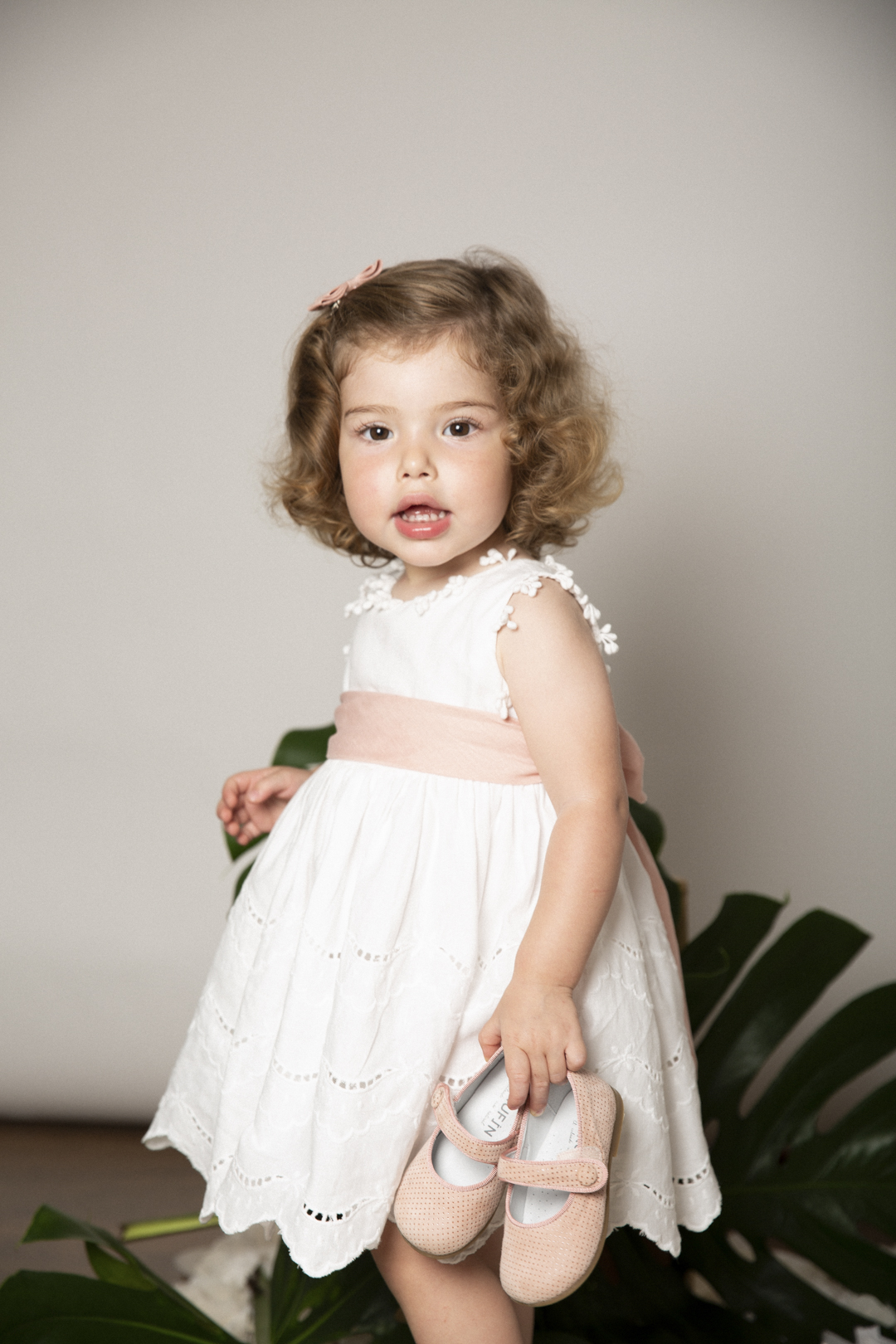 Niña con vestido blanco sujetando sus zapatitos en estudio