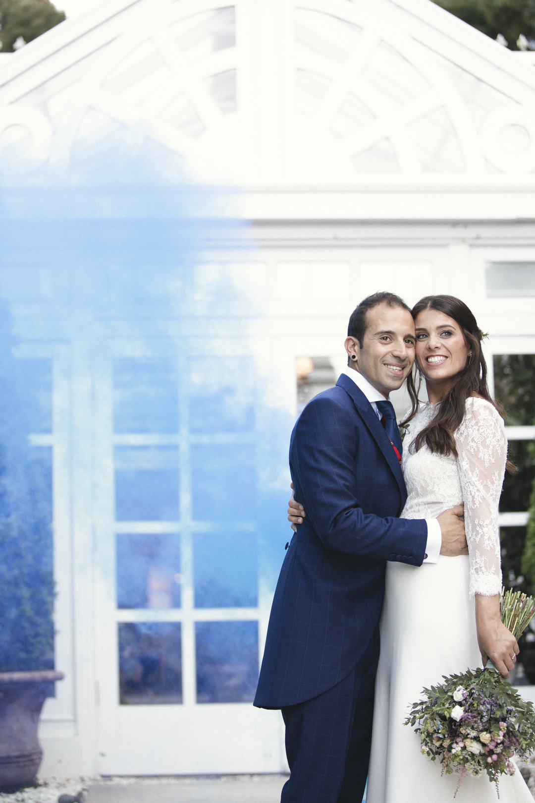Novios posando con humo de color frente a una fachada acristalada