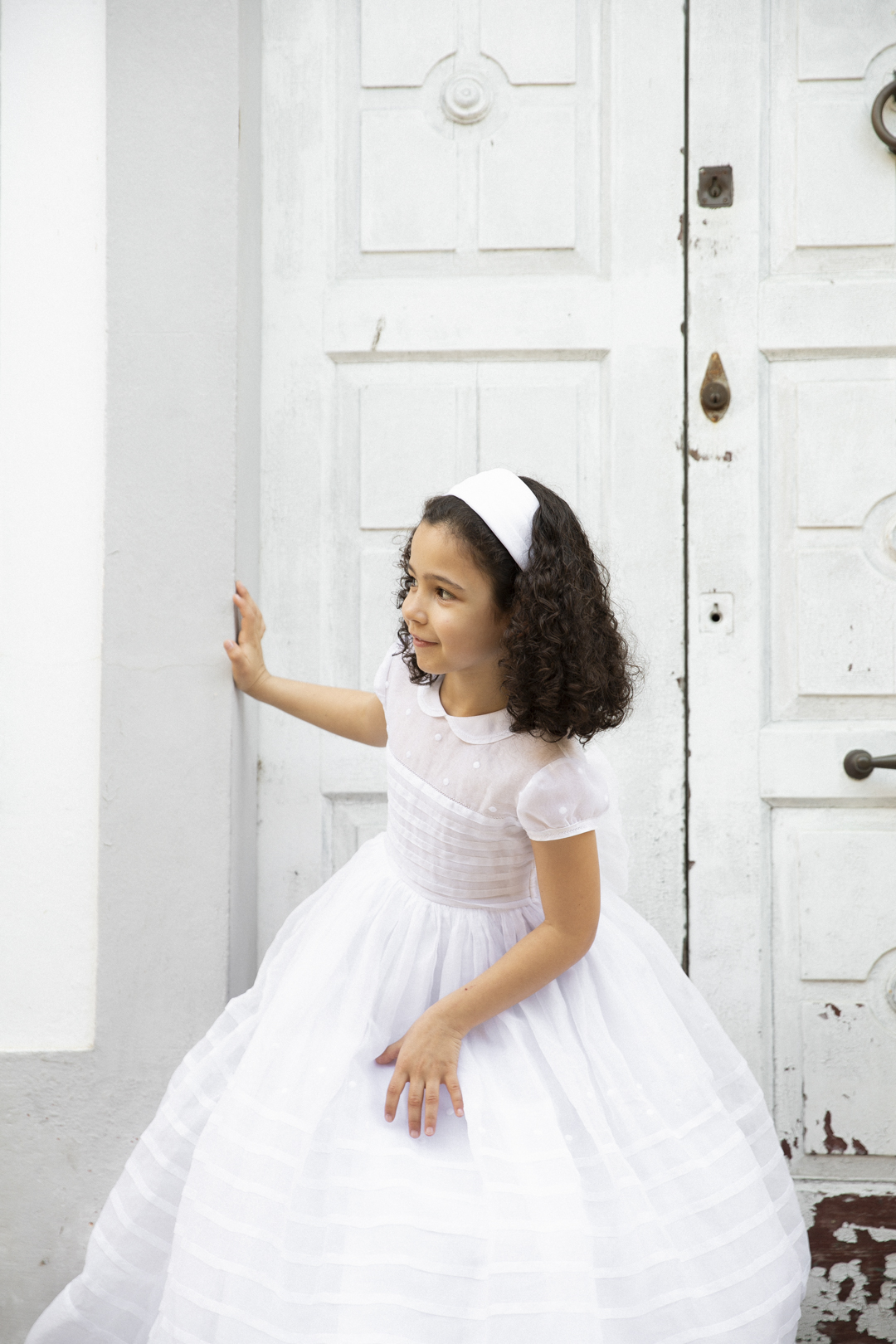 Niña con vestido de comunión frente a una gran puerta blanca