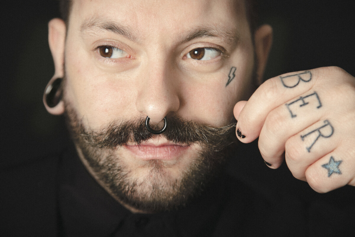 Retrato de barbero tatuado mirando a cámara mientras se atusa el bigote